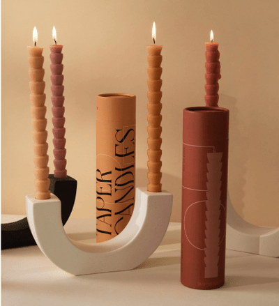 Four lit, ribbed taper candles in two modern U-shaped holders are displayed with cylindrical candle packaging against a beige background.