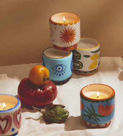 Four lit candles in colorful, patterned holders are arranged on a cloth with a tomato, artichoke, and yellow cherry tomato.