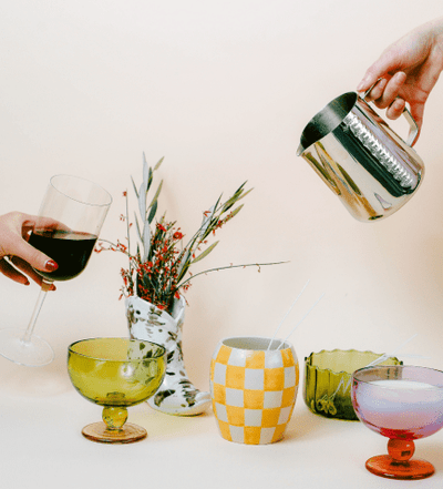 Two hands are shown: one holding a wine glass, the other pouring from a metal pitcher. Colorful glassware and a vase with flowers are arranged on a light surface.